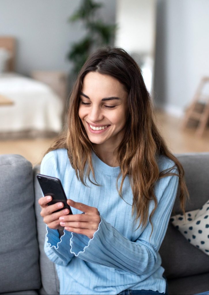 Cheerful young woman using mobile phone while sitting on a couch at home. Communication.